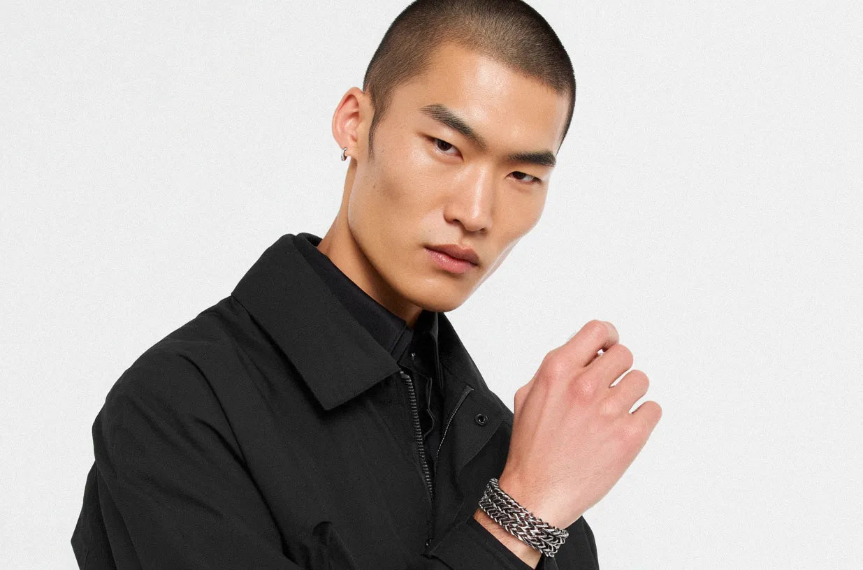 A man in a black collared shirt and jacket, featuring a textured Stainless Steel Bracelet on his left wrist.