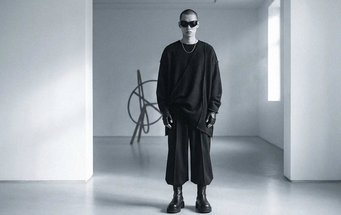 Asian male model wearing an avant-garde fashion outfit with an oversized black draped wool wrap top featuring raw exposed seams, cropped wide-leg trousers, chunky platform boots and fingerless leather gloves in a minimalist white gallery with an abstract sculpture in the background