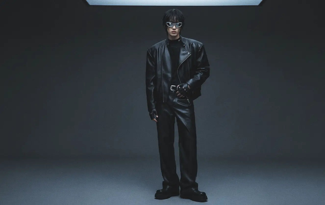 Asian male model in a full black avant-garde clothing look featuring an oversized faux leather biker jacket, wide-leg leather trousers and chunky platform boots under a dramatic overhead light