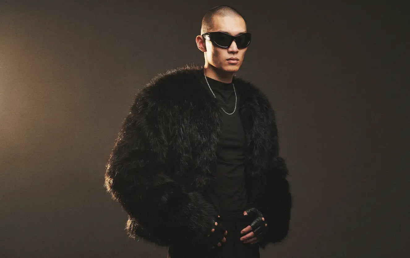 Asian male model wearing a dark avant-garde fashion outfit with an oversized black faux fur coat, black mock-neck top, silver chain and fingerless leather gloves against a warm-toned studio background