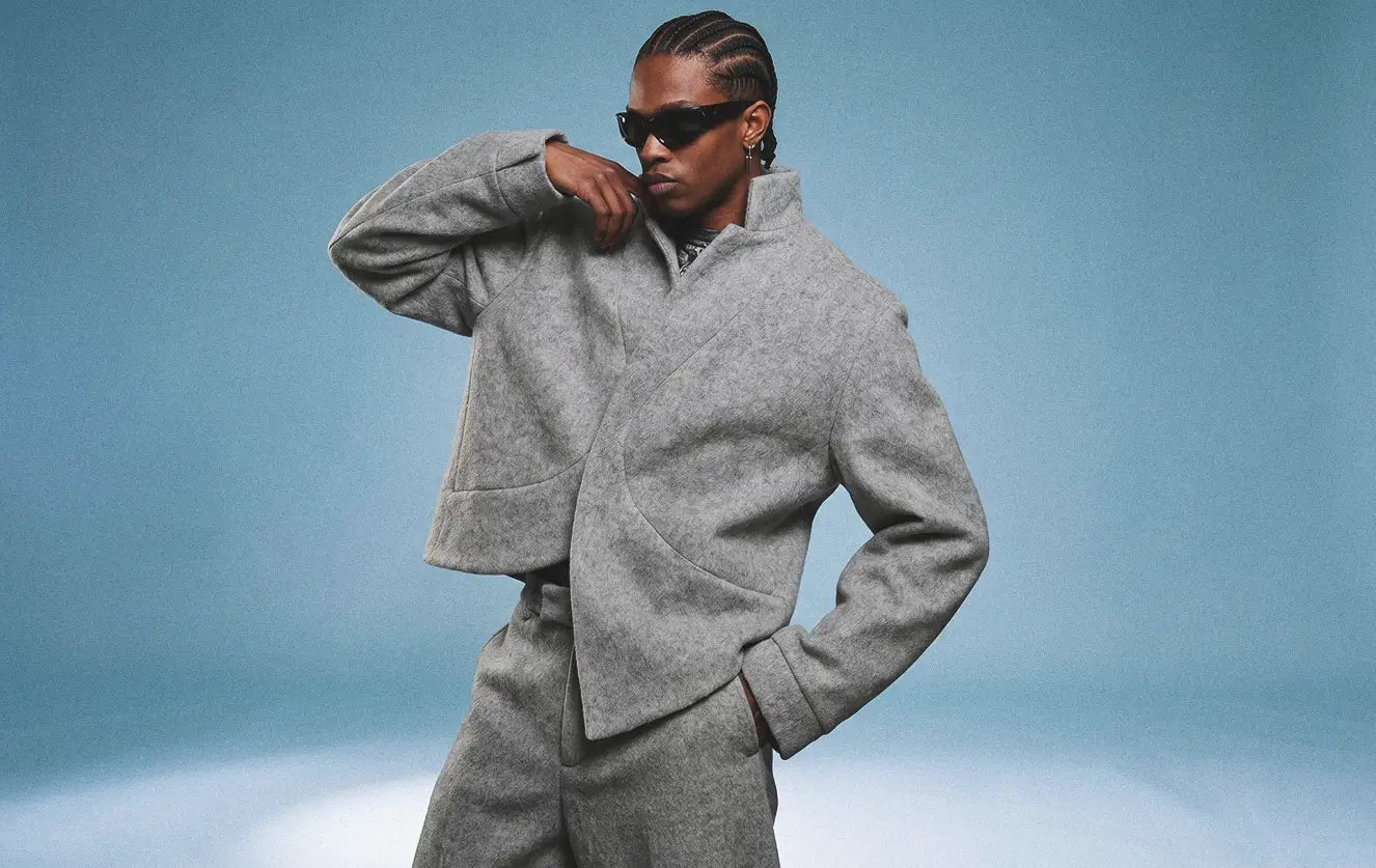 Black male model wearing an oversized grey wool felt wrap jacket with asymmetric curved closure and matching wide-leg grey wool trousers in an avant-garde fashion editorial studio shot