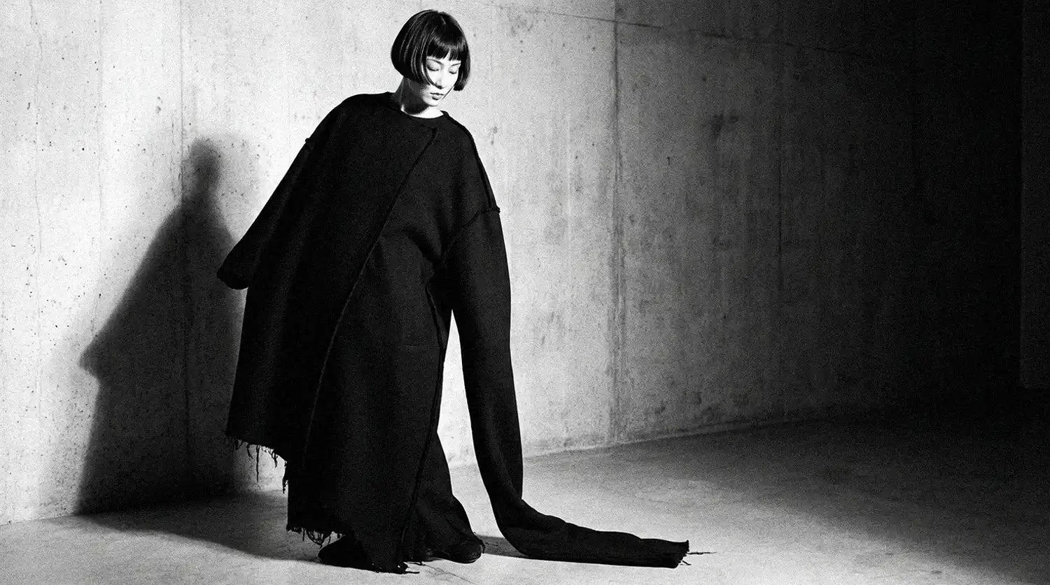 Japanese female model in oversized black wool coat with raw frayed hem and asymmetric seam against concrete wall, Yohji Yamamoto 1981 style