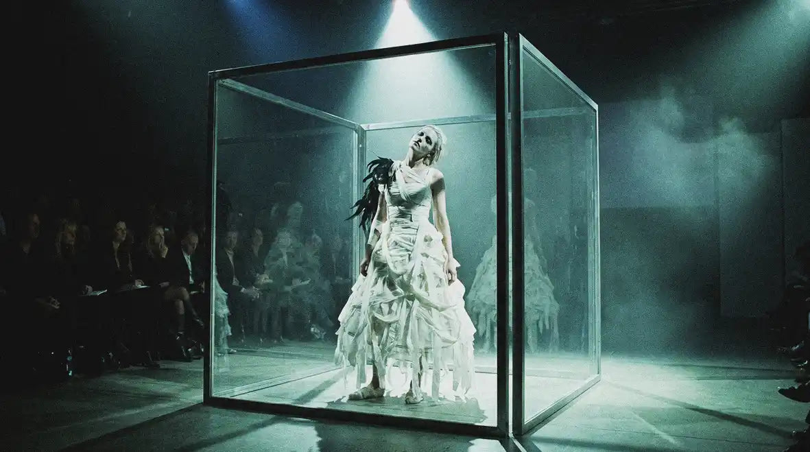 Theatrical avant-garde fashion show with model in white bandage dress and black feathers inside mirrored glass cube under dramatic spotlight