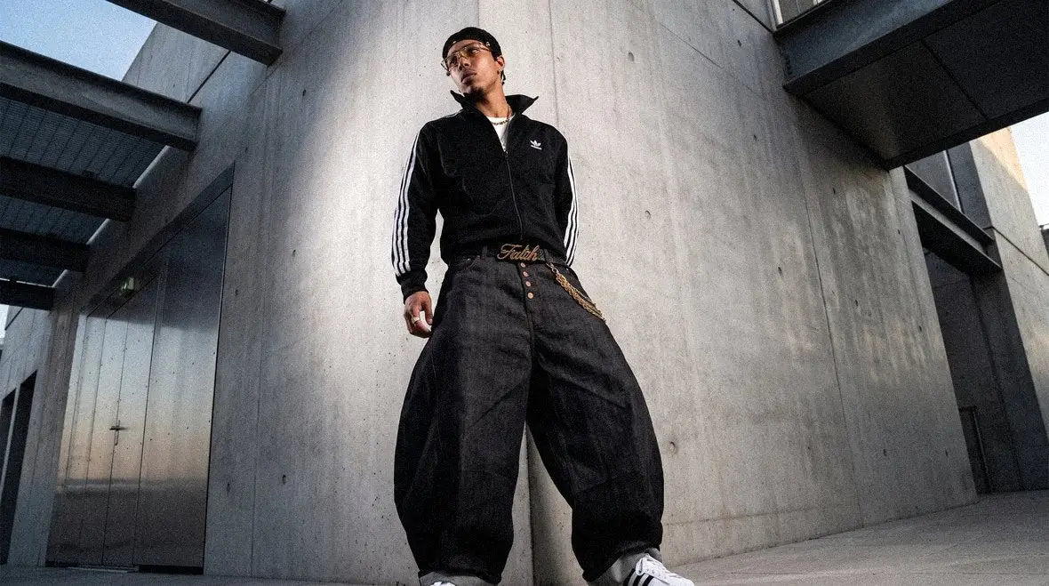 Man in a black track jacket and layered chain wears black barrel jeans with a statement belt buckle