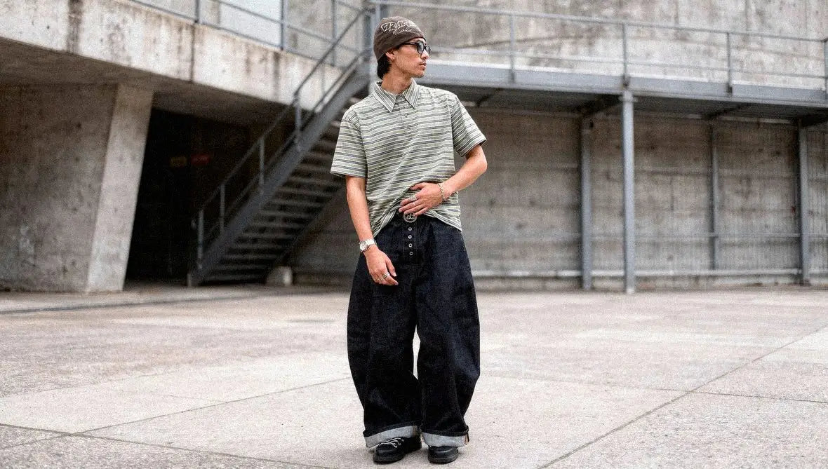 Man in a striped polo and beanie wears dark raw denim barrel jeans cuffed above black lace-up boots