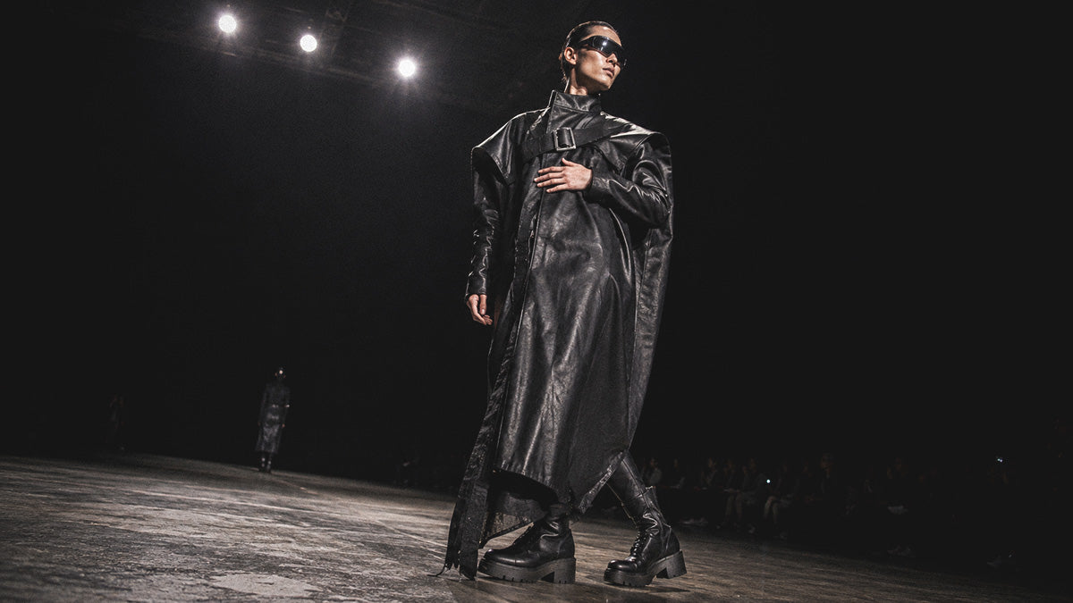 Model walking a dark runway in an oversized black leather trench coat, combat boots, and sunglasses for a cyberpunk techwear fashion show.