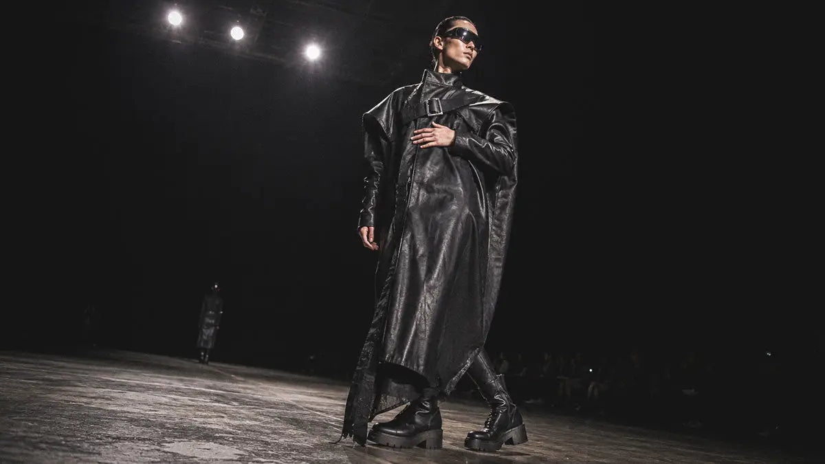 Model walking a dark runway in an oversized black leather trench coat, combat boots, and sunglasses for a cyberpunk techwear fashion show.
